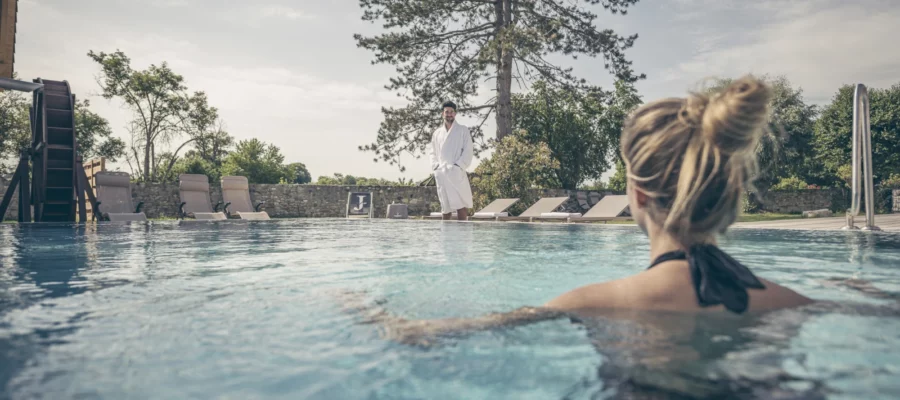 Entspannung am Außenpool im Spa © Jordans Untermühle Blick über einen Außenpool auf eine Person im Wasser mit hochgesteckten Haaren im Vordergrund. Im Hintergrund steht eine weitere Person im weißen Bademantel am Beckenrand, umgeben von Liegestühlen, Bäumen und einer Gartenanlage.