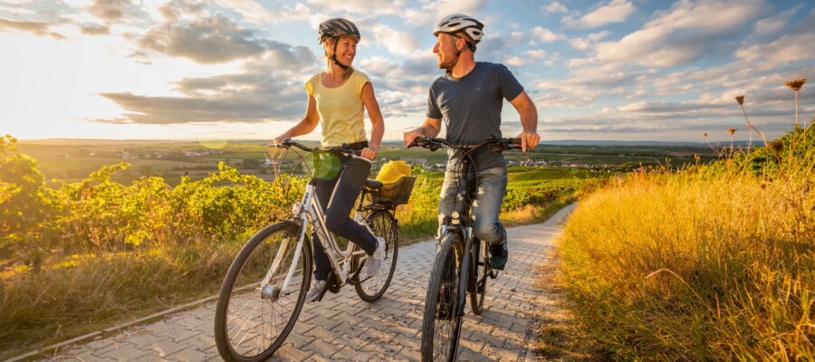 Radtour auf Weg durch Weinlandschaft, Gau-Odernheim, Petersberg Zwei Personen mit Fahrradhelmen fahren auf Fahrrädern einen gepflasterten Weg entlang, umgeben von Weinbergen und hohem Gras. Im Hintergrund sind Felder und eine weite Landschaft unter einem Himmel mit verstreuten Wolken im warmen Abendlicht sichtbar.