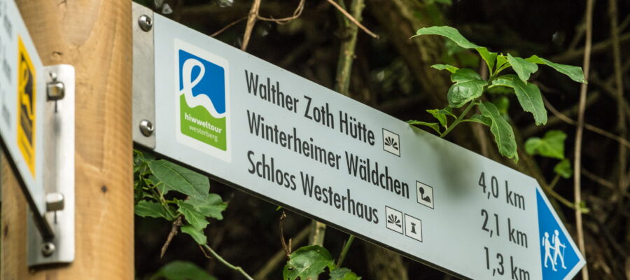 Beschilderung, © Dominik Ketz Nahaufnahme eines Wanderwegweisers an einem Holzpfosten in bewaldeter Umgebung. Das Schild der „hiwweltour westerberg“ weist auf „Walther Zoth Hütte“, „Winterheimer Wäldchen“ und „Schloss Westerhaus“ mit Entfernungen von 4,0 km, 2,1 km und 1,3 km hin.