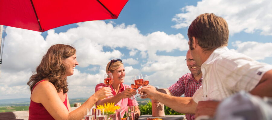 Gemeinsamer Genuss auf der Sonnenterrasse Vier Personen sitzen an einem Holztisch im Außenbereich und stoßen mit roséfarbenen Getränken an. Ein großer roter Sonnenschirm spendet Schatten, auf dem Tisch stehen Gläser und kleine Speisen, im Hintergrund sind Himmel und Landschaft zu sehen.