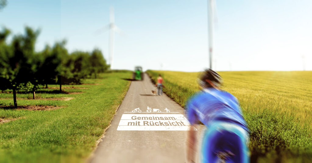 Bodenmarkierung Grafik Ein asphaltierter Weg verläuft zwischen Feldern und Bäumen. Im Vordergrund fährt eine unscharfe Person mit dem Fahrrad, auf dem Weg steht die Markierung „Gemeinsam mit Rücksicht“.