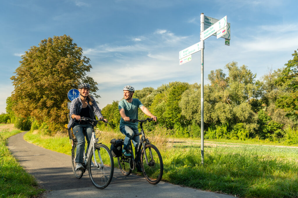 HBR-Beschilderung (3) Zwei Personen mit Fahrradhelmen fahren auf einem asphaltierten Radweg. Daneben steht ein Radwegweiser, im Hintergrund sind Bäume, Wiesen und blauer Himmel zu sehen.