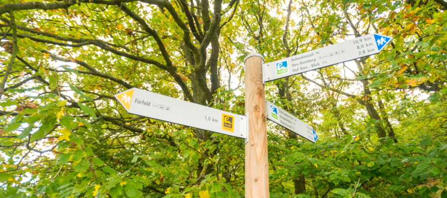 Beschilderung an der Hiwweltour Eichelberg, © Dominik Ketz Holzpfosten mit mehreren Wegweisern in einem grünen Wald. Die Schilder zeigen unter anderem Ziele wie Fürfeld, Robenkanzel, Neu-Bamberg und Hof Iben – Blick mit Entfernungsangaben.