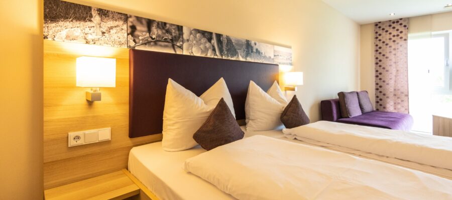 Weingut Meyerhof Helles Hotelzimmer mit zwei nebeneinanderstehenden Betten, weißen Kissen und violetten Zierkissen. Wandlampen, Holzverkleidung, Fotoleiste und ein violettes Sofa ergänzen die Einrichtung.