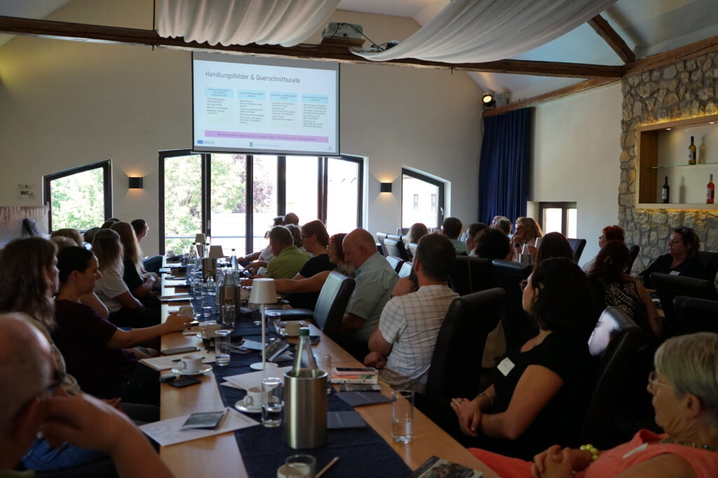 Präsentation vor einer großen Besuchergruppe Eine große Gruppe sitzt an langen Tischen in einem hellen Veranstaltungsraum und blickt auf eine Präsentationsleinwand. Sichtbar sind Getränke, Unterlagen, Holzbalken, Stoffbahnen und eine Natursteinwand.
