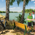 Neuer Radweg stärkt die Qualitätsradregion Zwei Personen stehen unter Birken an einem Gewässer und betrachten eine aufgefaltete Karte. Neben einer Holzbank stehen mehrere Fahrräder mit Helmen. Links ist die Grafik „2030+“ eingeblendet.