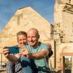 Digitale Sprechstunde: Praxisnah. Persönlich. Für Ihren Betrieb. Zwei Personen sitzen vor einer historischen Steinruine und fotografieren sich mit einem Smartphone. Links ist auf einer violetten Fläche deutlich der Schriftzug „2030+“ eingeblendet.