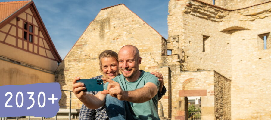 2030-plus_B2B_9.1_Smart_Destination_Rheinhessen Zwei Personen sitzen vor einer historischen Steinruine und fotografieren sich mit einem Smartphone. Links ist auf einer violetten Fläche deutlich der Schriftzug „2030+“ eingeblendet.