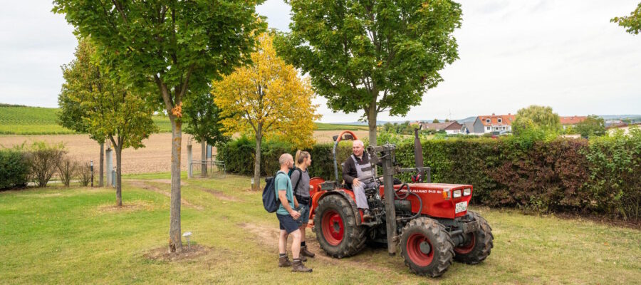 Siefersheim - Reisemobilstellplatz Weingut Sommer Zwei Personen mit Wanderrucksäcken stehen bei einem Mann auf einem roten Traktor. Die Szene befindet sich auf einer Grünfläche mit Bäumen; im Hintergrund erstrecken sich Felder, Weinberge und eine Ortschaft.