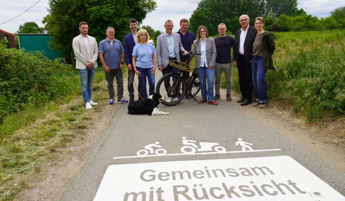 Zehn Personen stehen mit einem Fahrrad und einem liegenden Hund auf einem asphaltierten Weg. Im Vordergrund zeigt die Bodenmarkierung „Gemeinsam mit Rücksicht“ Symbole für Radverkehr, Landwirtschaft und Fußverkehr.