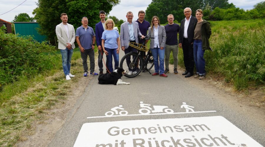 Zehn Personen stehen mit einem Fahrrad und einem liegenden Hund auf einem asphaltierten Weg. Im Vordergrund zeigt die Bodenmarkierung „Gemeinsam mit Rücksicht“ Symbole für Radverkehr, Landwirtschaft und Fußverkehr.